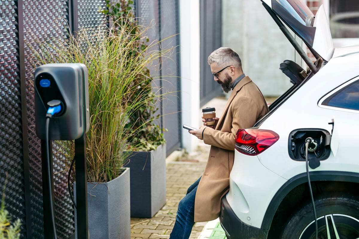 Kasko parim lisakaitse - soliidne härrasmees laetava valge elektriauto vastu toetumas, laadimisjaam selle juures.